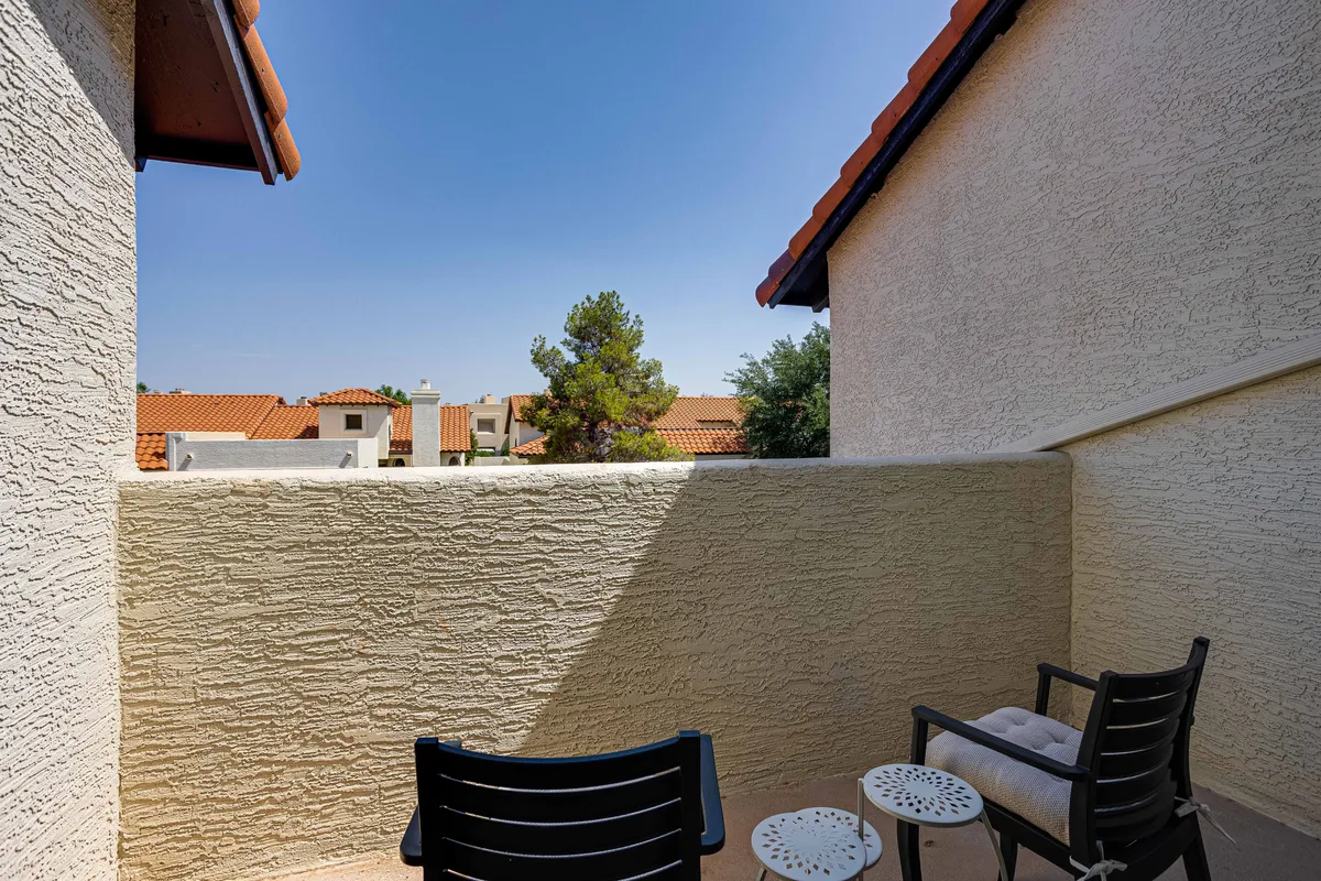 An exterior shot showing a private front courtyard space with two black chairs and a small round table surrounded by greenery, with red-tiled roofs in the background. The scene embodies relaxation and privacy at the threshold of the home.