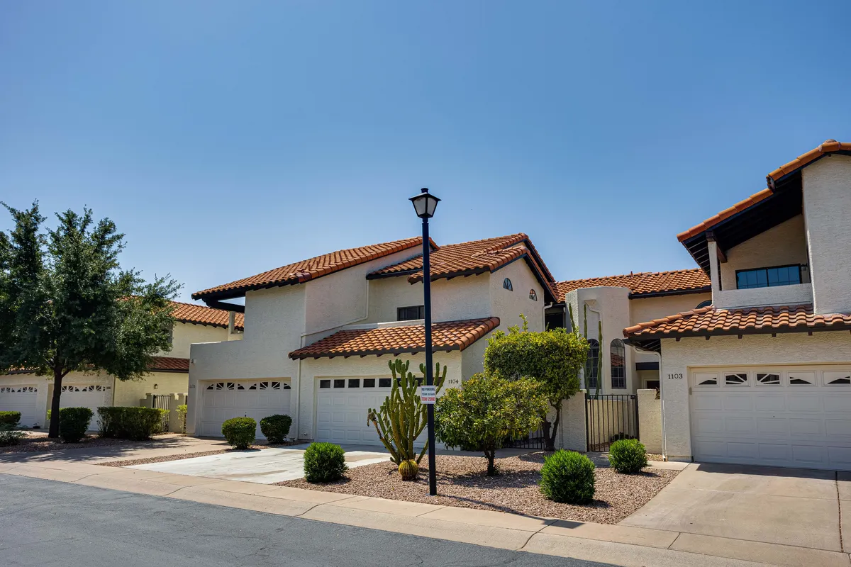 This image captures the architectural features of the building surrounded by lush landscaping with cacti and shrubs, highlighted by a sunny, inviting atmosphere. The street lamp and open walkway evoke a sense of community and leisurely outdoor living.