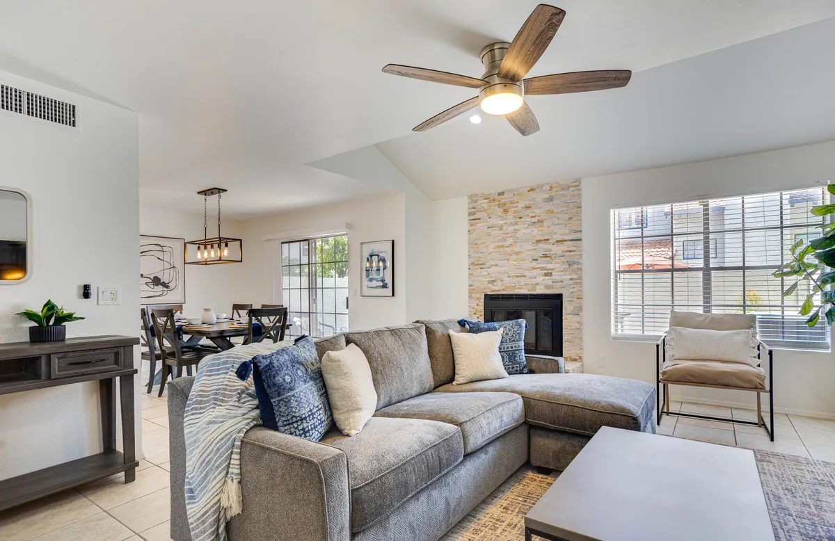 This image features a welcoming open-concept living area with a sectional sofa centered around a striking stone fireplace. The dining area is visible in the background, emphasizing the seamless flow between spaces. Large windows allow for abundant natural light, creating a bright and cozy environment.