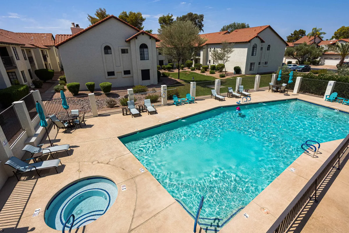 A serene outdoor scene showing a sparkling swimming pool and adjacent hot tub, surrounded by lush greenery and comfortable lounge chairs. The background features attractive residential buildings, and the overall mood is leisurely and tranquil, ideal for sunbathing, relaxing, or socializing.