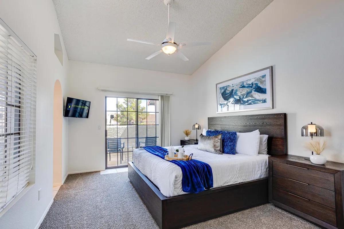 A calm, light-filled bedroom featuring a neatly made bed, a window offering a pleasant view, a nightstand with a lamp, and tasteful artwork on the wall. The space feels tranquil and inviting, perfect for restful sleep or quiet moments.