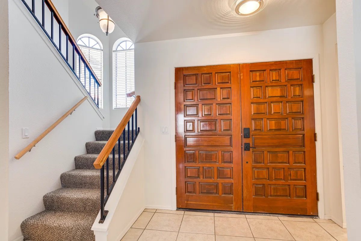 This image captures large wooden front doors opening into a well-lit entryway, with a staircase and windows allowing plenty of natural light to stream in. The welcoming foyer provides a first impression of spaciousness and ease as one enters the home.